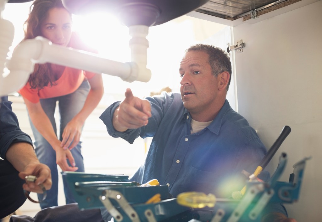 Plumber pointing to a pipe under a sink while explaining a repair to a homeowner.